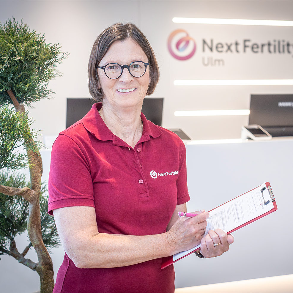 Eine Mitarbeiterin von Next Fertility Ulm steht am Empfang und schreibt etwas in ein Formular.a